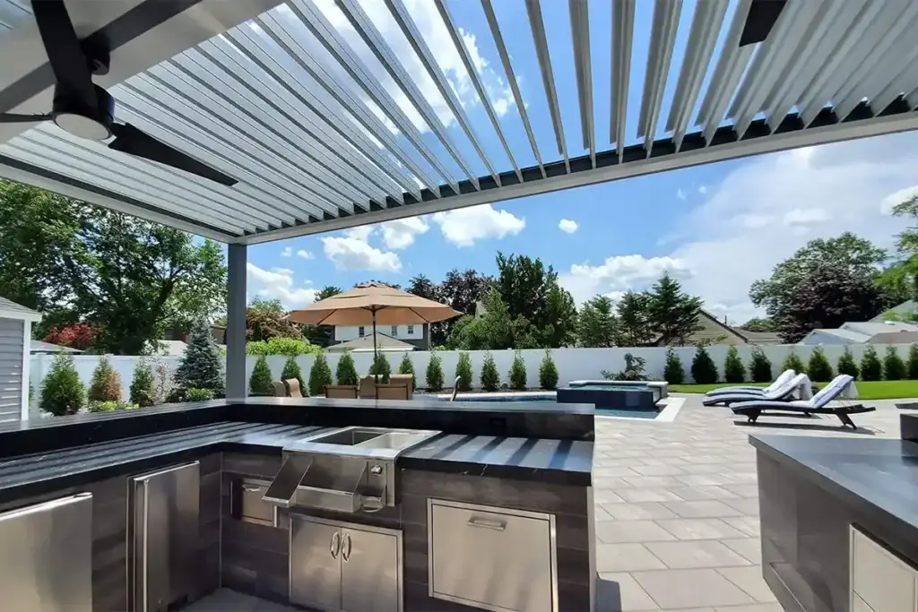 Outdoor stainless steel kitchen under a slatted pergola overlooking a pool area with lounge chairs and a tan umbrella across a manicured yard.