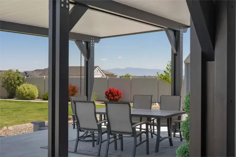 Outdoor patio under a dark pergola with a glass-top table and gray chairs, in a yard with green lawn and distant mountains.