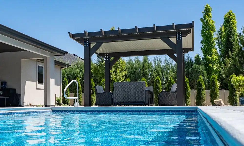 Sunny backyard with a rectangular blue pool and a dark wooden pergola patio featuring wicker seating and a coffee table behind the pool edge.