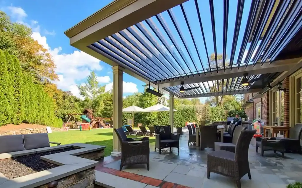 Outdoor patio under a slatted pergola with wicker chairs and a table, overlooking a green yard with a playground and umbrellas in the distance