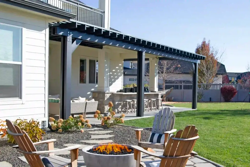 Backyard patio with a lit fire pit, wooden Adirondack chairs, and a metal-framed pergola under a sunny blue sky.