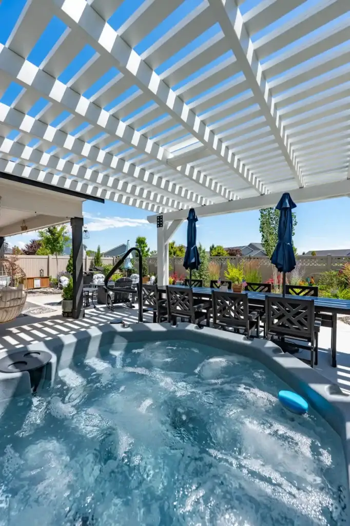 Outdoor backyard patio with a white slatted pergola over a bubbling hot tub and blue sky above.