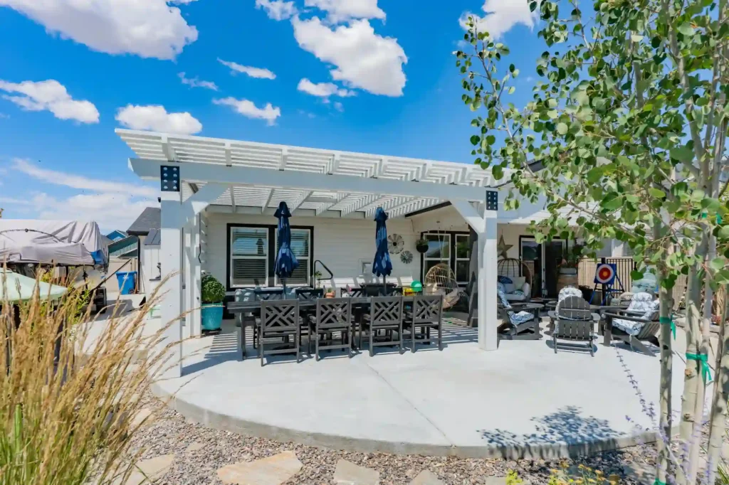 White pergola over a gray outdoor dining area with a long table and blue umbrellas in a sunny backyard.