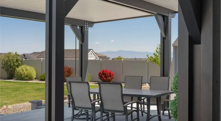 Outdoor patio under a dark pergola with a glass-top table and gray chairs, in a yard with green lawn and distant mountains.