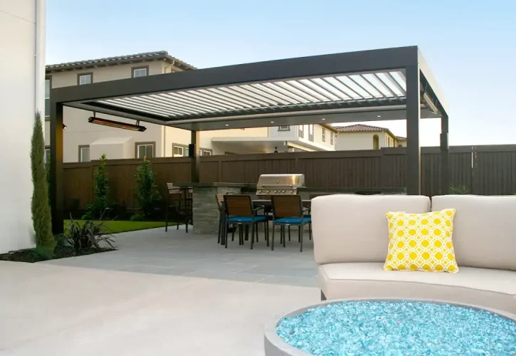 Backyard patio under a black pergola with an outdoor kitchen, dining table, and beige sectional sofa with a yellow pillow; a blue glass fire pit in the foreground.
