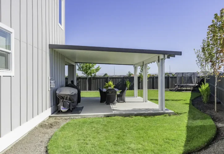 Backyard with a covered patio attached to a gray house, featuring a dining table, chairs, and a grill; trampoline visible in the distant yard.