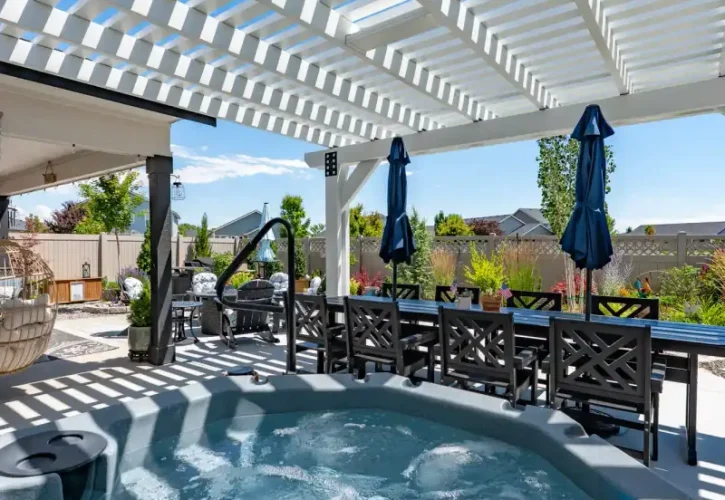 Outdoor backyard patio with a white slatted pergola over a bubbling hot tub and blue sky above.