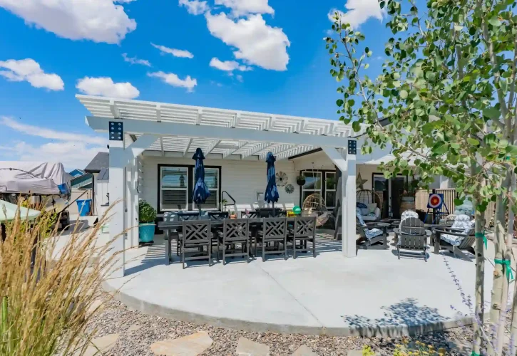 White pergola over a gray outdoor dining area with a long table and blue umbrellas in a sunny backyard.