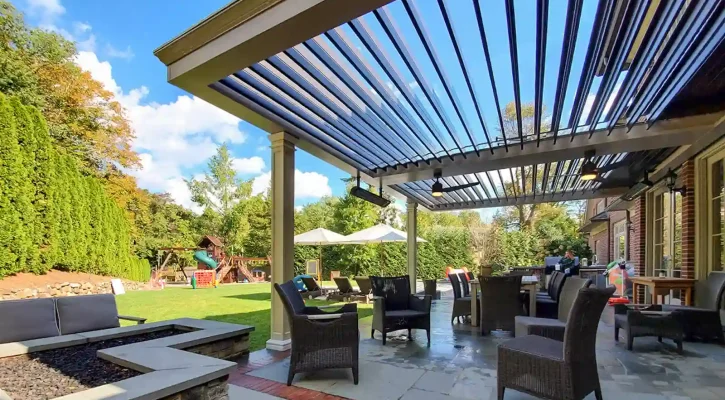 Outdoor patio under a slatted pergola with wicker chairs and a table, overlooking a green yard with a playground and umbrellas in the distance